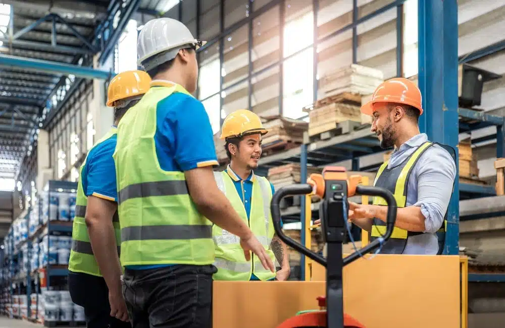 Warehouse workers in safety gear discussing lean training methods near forklift