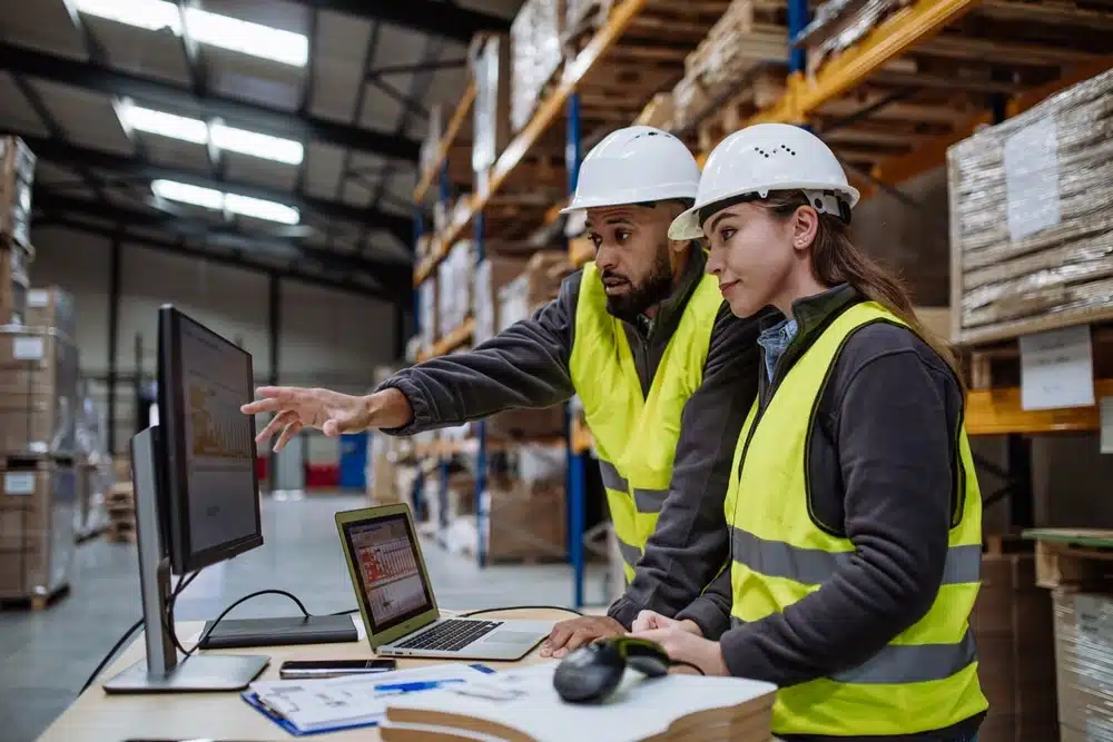 Two warehouse workers in safety gear using inventory management software on computer