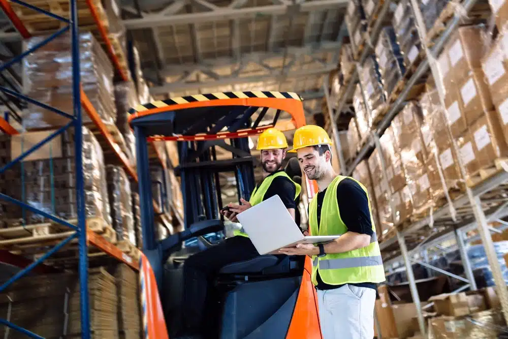 Two warehouse workers in safety gear reviewing barcode inventory data near forklift