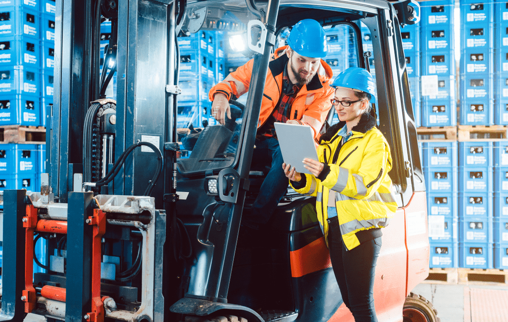 warehouse worker on a forklift talking to another warehouse worker holding a clipboard in a beverage distribution warehouse